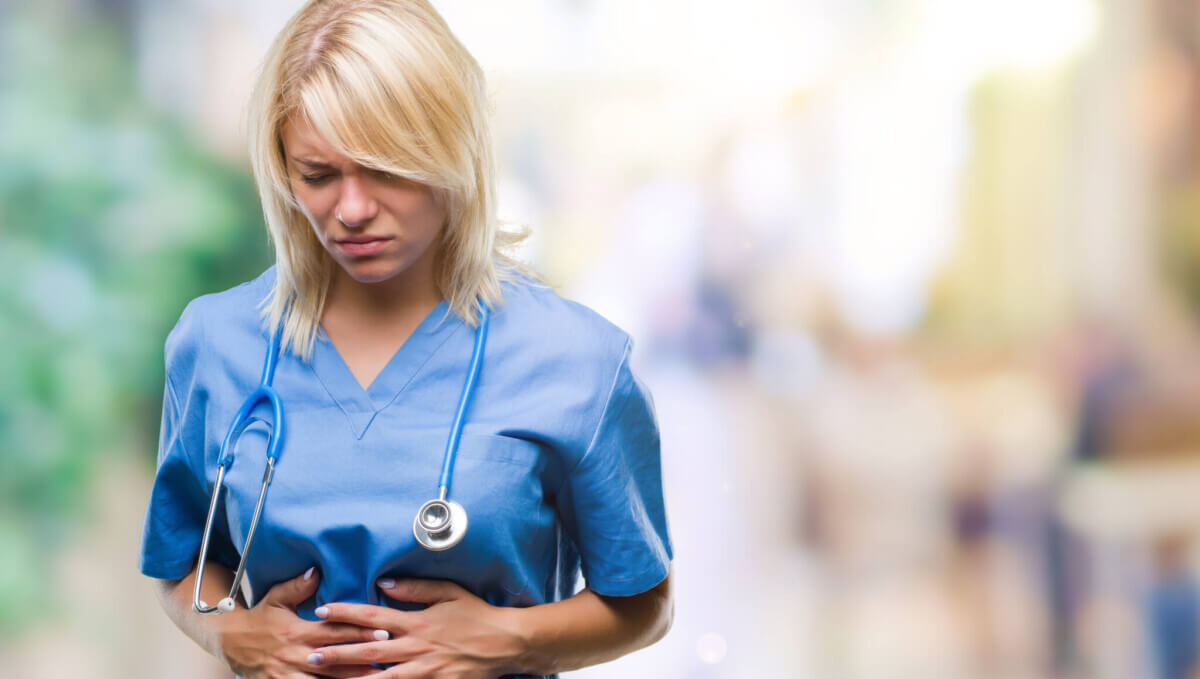 Young beautiful blonde doctor woman wearing medical uniform over isolated background with hand on stomach because nausea, painful disease feeling unwell. Ache concept.