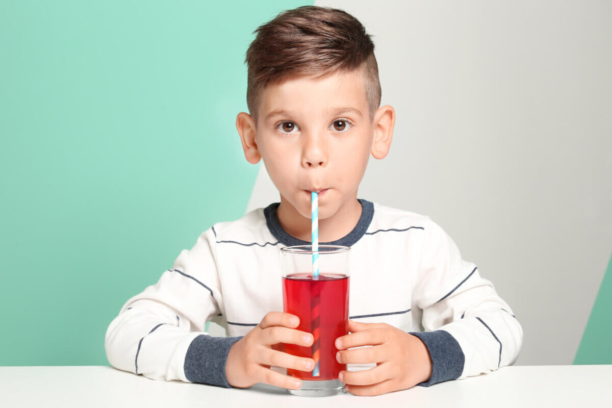 Cute little boy drinking juice while sitting at table, on color background