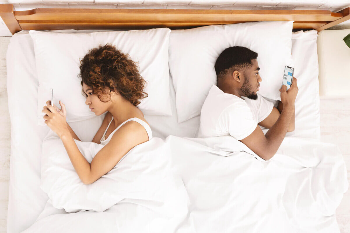 Millennial couple lying on bed back to back with smartphones