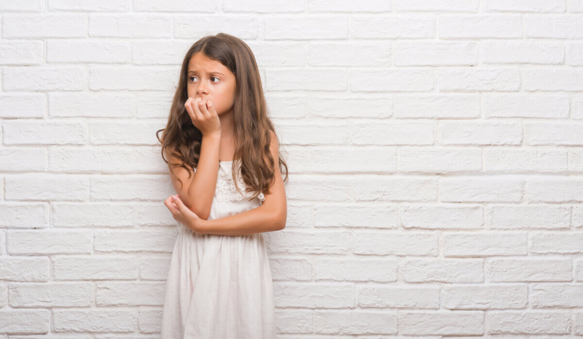 Young girl over white brick wall looking stressed and nervous with hands on mouth biting nails. Anxiety problem.