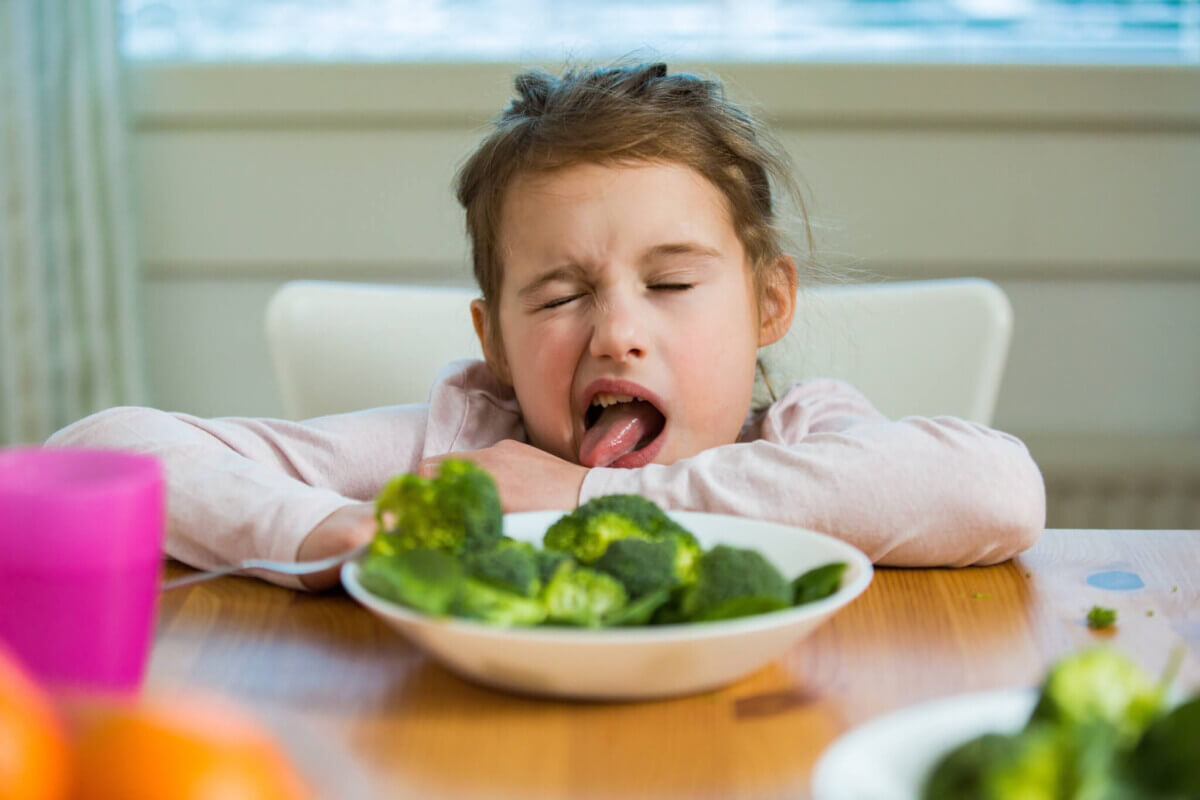 Cute girl eating spinach and broccoli at the table. Child doesn't want to eat, refuses eating, making faces. Healthy food concept.
