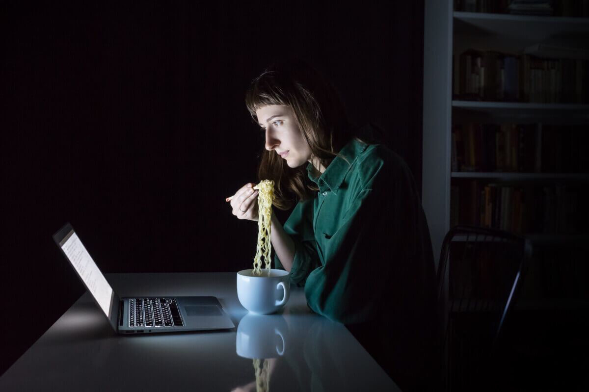 Young female at laptop computer eats instant ramen noodles late in the evening. Woman working or studying online overtime at night has fast dinner - concept of unhealthy junk food at workplace