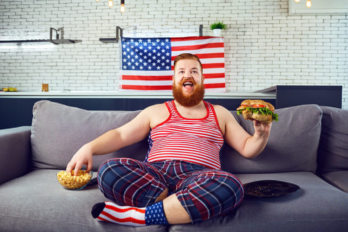Overweight funny man eating a burger, snacking while sitting on the couch