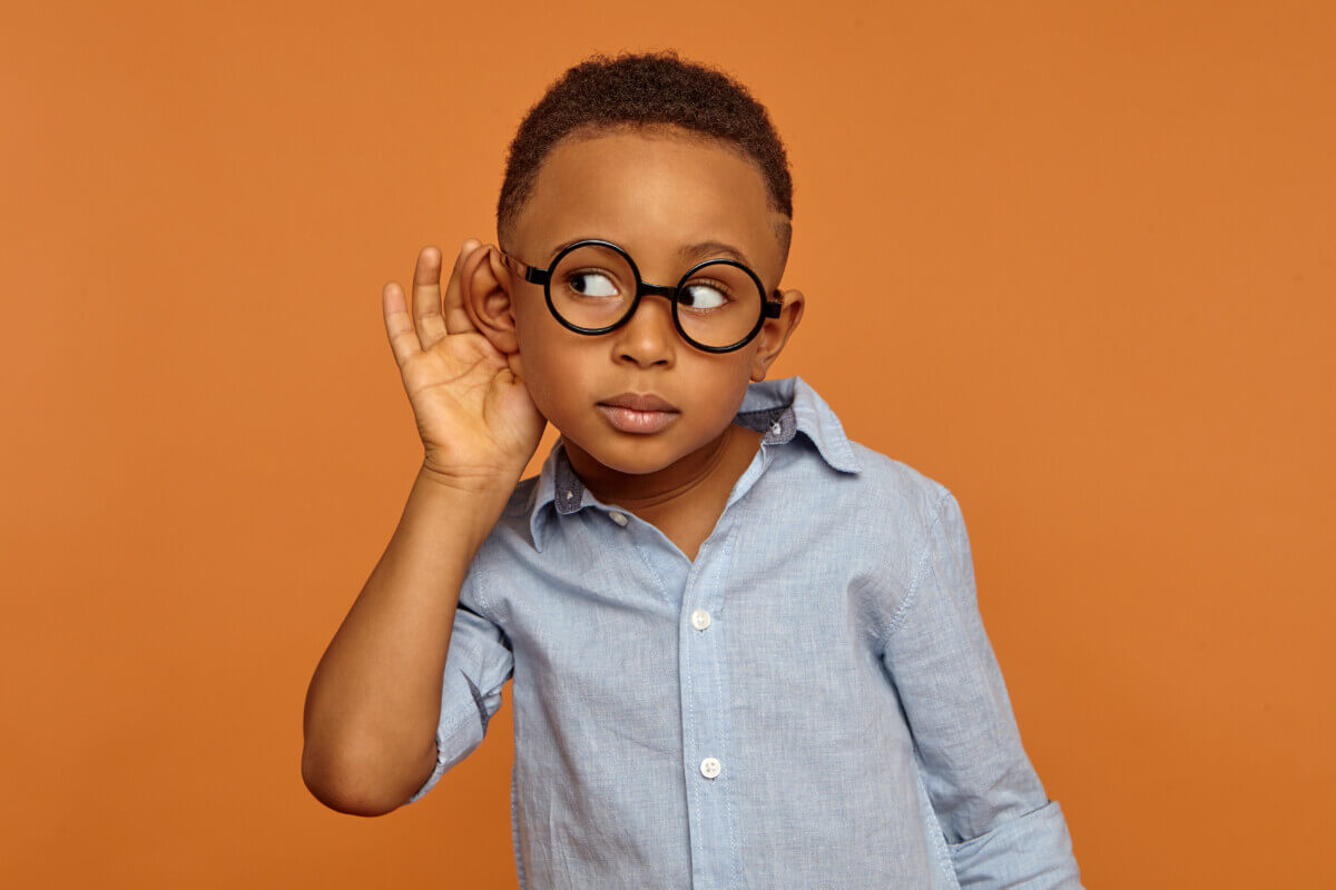I want to hear everything. Little boy wearing round spectacles and neat shirt having curious facial expression while spying, overhearing, holding hand on his ear
