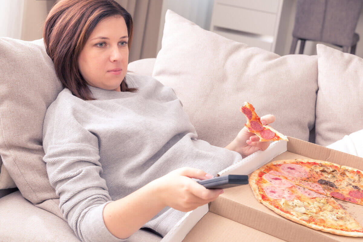 Surprised woman eating pizza and watching TV with remote control at home, warm tone