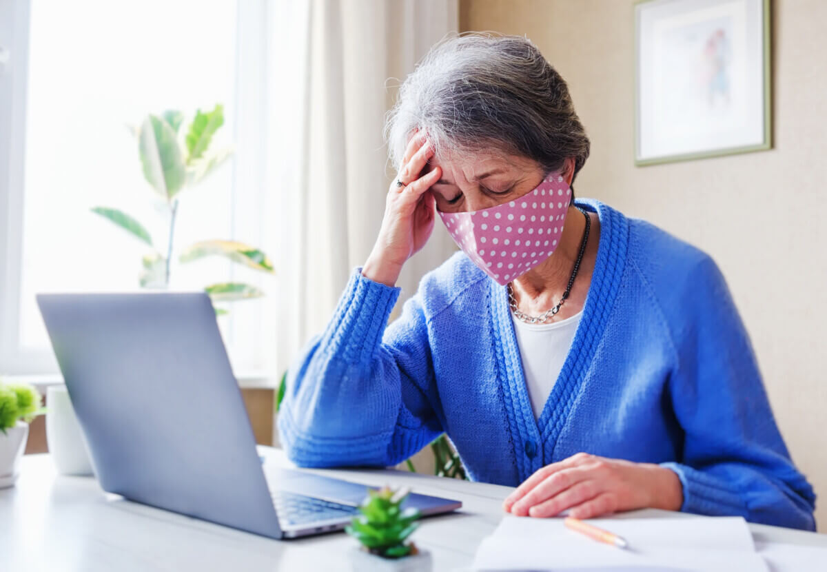 An elderly woman in a mask with a laptop is experiencing stress and headache amid the coronavirus pandemic - A pensioner wearing a face mask uses a computer to search for information on the Internet
